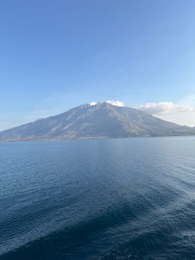 The image captures a wide-angle view of a mountainous coastal landscape near Palaiopoli, Samothrace, Greece. The foreground is dominated by a large body of deep blue water exhibiting subtle ripples across its surface. In the midground, a substantial, rugged mountain range, likely Mount Saos, extends horizontally across the frame, rising directly from the coastline. Its slopes display a mix of rocky outcroppings and sparse, dull-toned vegetation. Small, white cumulus clouds are observed near the upper peaks on the right side of the mountain. A faint, small white structure is barely visible on the distant shoreline at the mountain's base. The background consists of a clear, light blue sky with minimal cloud cover, primarily concentrated around the mountain peaks. No individuals or vessels are present in the depicted scene.