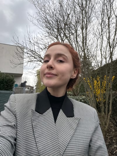 This image depicts a residential outdoor scene, located in the city of Eschau, France. A young adult female, with reddish-brown hair styled back and fair skin, is centrally positioned, looking directly at the camera with a subtle, closed-mouth smile. She wears a black turtleneck sweater beneath a black and white small-checked blazer featuring broad black lapels. A small stud earring is visible in her left earlobe. The background behind her is composed of multiple bare deciduous trees on the right and center, indicative of late winter or early spring. To the far right, a shrub with bright yellow flowers is visible. On the left side of the frame, a modern, multi-story white building is partially visible, behind which a grey structure and a black silhouette resembling a cat are observed. The sky above is overcast.