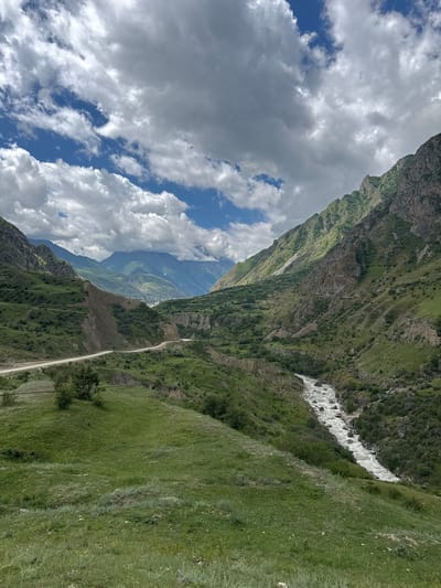 This is a landscape photograph taken in Verkhnyaya Balkariya, Russia. The image depicts a dramatic mountain valley with steep, green-covered slopes. A winding dirt road is visible, running along the side of one of the slopes. A fast-flowing river with white, turbulent water carves through the valley floor, surrounded by rocks. The sky is partially cloudy, with patches of blue sky showing through large, white, fluffy clouds. The time of day appears to be daytime, likely late morning or early afternoon, given the bright lighting and the shadows cast by the clouds. The overall impression is one of rugged natural beauty and vastness, with a sense of remoteness conveyed by the mountainous terrain and the solitary road. There is no visible text in the image.