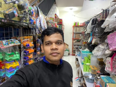 The media file is a selfie taken by a man in a store. The man is wearing a black jacket over a blue shirt and is looking directly at the camera. His face is partially visible due to the image being rotated. The background is filled with shelves displaying various items, including kitchenware, plastic containers, and toys.
In the foreground, on the left side, there are pots and pans wrapped in plastic. To the right of these, there are shelves with kitchen utensils like spatulas and ladles. Further to the right, shelves are stocked with colorful toys, including toy trucks and stacking toys, some of which are packaged in clear plastic. There are also various bottled liquids, possibly cleaning supplies or beverages, on some shelves. The setting appears to be a retail store in Lamteumen Barat, Indonesia. The lighting suggests it could be daytime, though no specific weather details are apparent. The man's expression is neutral. Visible text includes "ENGINEERING TRUCK" on a toy packaging.