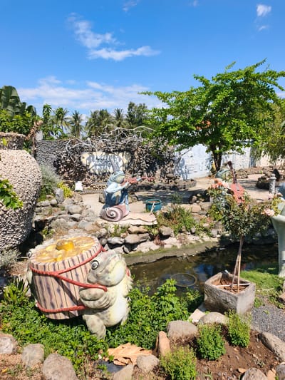 This image captures a whimsical garden scene in Tuy An Nam, Vietnam, featuring a pond, lush greenery, and quirky frog sculptures. In the foreground, a large, light-colored frog sculpture is depicted holding a wooden barrel filled with yellow items, possibly fruit, with red ropes securing it. The frog is positioned on a bed of vibrant green grass, surrounded by several large, natural rocks.
To the left, a large, textured, cream-colored pot is partially visible, adorned with leaves. Behind the barrel-carrying frog, a small pond reflects the blue sky and white clouds. The pond is bordered by rocks and more green vegetation.
Further back, another frog sculpture, this one sitting on a snail shell, is shown playing a stringed instrument. Its attire suggests it's wearing a hat and a blue top. Adjacent to this, a tree with dense green foliage provides shade.
The background features a wall constructed from dark stones, with decorative white artistic elements and a sign with Vietnamese text. Palm trees and other tropical foliage are visible beyond the wall, under a clear blue sky dotted with fluffy white clouds. The overall atmosphere is one of a creatively decorated outdoor space, likely a garden or park attraction, with a focus on playful frog imagery.
