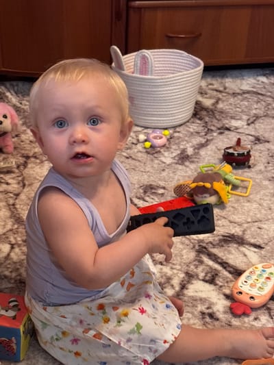 A young Caucasian child with blond hair and blue eyes is sitting on a patterned rug in a room, surrounded by toys. The child is wearing a light purple tank top and a white skirt with a floral pattern. The child is holding a black object with red accents in their hands. Nearby are several toys, including a pink stuffed animal, a toy phone, a drum, and other colorful plastic toys. In the background, there is a wooden dresser and a white woven basket with pink bunny ears. The image appears to be taken indoors.
