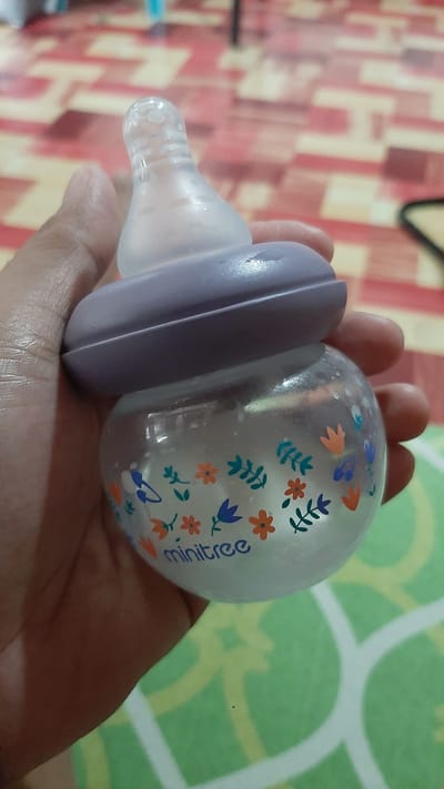 A person is holding a clear baby bottle with a purple ring and a nipple. The bottle has a floral pattern with orange and blue flowers and green leaves. The word "minitree" is visible in blue lettering on the bottle. The bottle appears to contain a white liquid. The background is out of focus, showing a patterned floor and a green surface with a white pattern. The image appears to have been taken indoors.