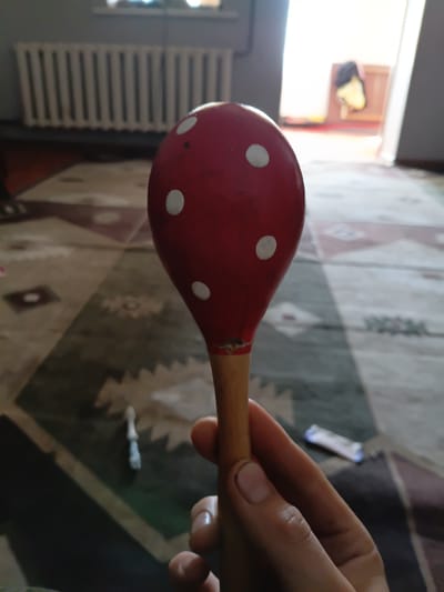 A red maraca with white polka dots is held in a person's hand. The maraca has a wooden handle and a round top painted red with white spots. In the background, a radiator is visible on a wall, and a doorway leads to another room where clothes are hanging. The scene appears to be indoors, possibly in a residential setting.