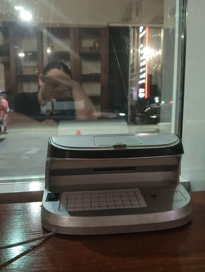 A person is reflected in a window, holding their hand to their face. They are wearing a black t-shirt. To the left, a person in pink is visible. In the foreground, a silver and black electronic device, possibly a scanner or printer, sits on a wooden surface. The device has a grid etched onto its surface. Behind the device, the window reflects the interior of a room with shelves and lighting, as well as an exterior view of a street with a neon sign that reads "ART CAFE" in vertical text. The scene appears to be set indoors, possibly at night, given the reflections and lighting. The overall atmosphere is somewhat blurry due to the reflections and the nature of the window.