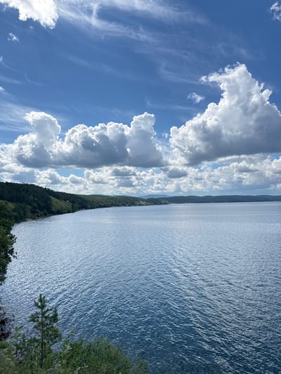 The image depicts a serene, wide body of water, likely a lake or reservoir, with a distant, tree-covered shoreline under a bright blue sky filled with dramatic white cumulus clouds and wispy cirrus formations. The water's surface is gently rippled, reflecting the sky's light and creating a textured, shimmering effect. The left side of the frame features a rocky, vegetated bank with a small evergreen sapling in the foreground. The shoreline stretches across the mid-ground, densely populated with evergreen trees that form a natural, rolling landscape. In the far distance, a faint hazy line of hills can be seen, suggesting a vast, open environment. There are no people or visible man-made structures in the immediate foreground or on the water. The overall impression is one of natural beauty and tranquility, consistent with a location like Dom Otdykha Zolotoy Plyazh, Russia, known for its natural landscapes.