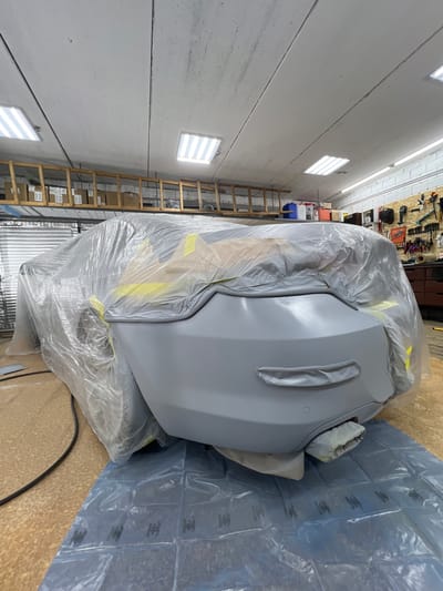 The image shows a car part, likely a bumper, covered in grey primer and plastic wrap. The part is resting on a blue plastic sheet on a wooden floor. In the background, there are shelves with tools and supplies, a wooden ladder leaning against a wall, and fluorescent lights on the ceiling. The setting appears to be a workshop or garage, possibly for car repair or painting. The overall impression is one of preparation for painting or a similar automotive process. There is no visible text in the image.