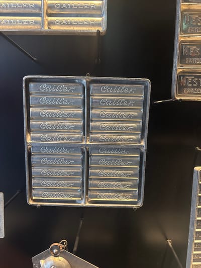The image depicts a display of vintage metal chocolate molds, likely at the Lindt Home of Chocolate museum in Kilchberg (ZH), Switzerland.  The molds, branded with the name "Cailler," are rectangular with multiple cylindrical depressions, presumably to create chocolate bars. Several similar mold types surround the primary Cailler mold, including one branded Nestle. The molds are mounted on a dark, presumably black, background. The scene is well-lit, capturing details in the metal surfaces.