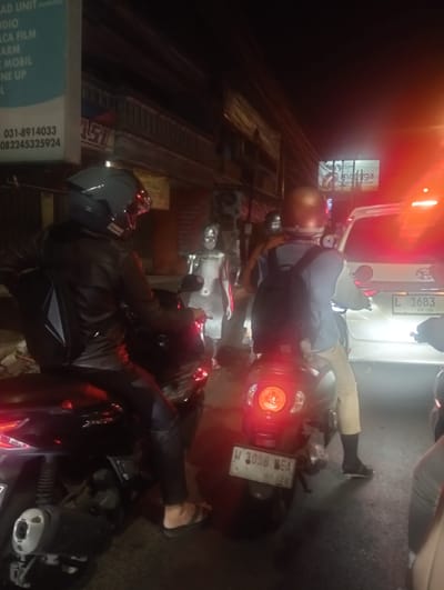 The image depicts a street scene at night, likely in the city of Calukan RT 1 RW 5 Keboansikep, Indonesia, based on visual cues like the license plate format and ambient lighting. In the foreground, two individuals are seated on motorcycles. The motorcycle on the left is black and ridden by a person wearing a black leather jacket and a dark helmet. A black backpack is visible on their back. The motorcycle on the right is also black, and the rider wears a brown helmet, a blue jacket, and light-colored pants. They also have a black backpack. To the right of the motorcycles, a white car is visible, with its license plate reading "L 1683 10-30". The background shows buildings and signs illuminated by streetlights and vehicle lights. The overall atmosphere suggests traffic congestion on a city street at night.