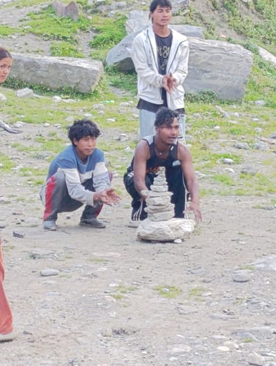 In the city of Marhi, India, three young men are gathered in an outdoor setting. One man, wearing a white jacket over a dark t-shirt and light-colored pants, stands with his hands clasped. To his right, another man kneels, stacking stones into a cairn. He is wearing a dark tank top and dark pants. In front of him, a third man is crouched, also with his hands open and facing forward. He is wearing a blue and white striped long-sleeved shirt and dark pants with red accents. The ground is covered in dirt and rocks, with patches of green grass visible. Large rocks are scattered throughout the background.