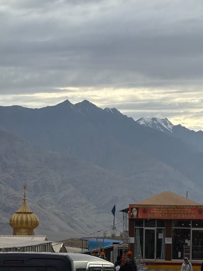 This is a vertical, eye-level shot of a mountainous landscape with a building in the foreground. The sky is overcast with streaks of light suggesting either sunrise or sunset. The mountains are rugged and appear to be covered in snow in some areas. In the foreground, a building with a sign that reads "GURUDWARA SHRI PATHAR SA" and "RECEPTION" is visible. The building has large glass windows. Several people are gathered near the building, some wearing turbans, suggesting a religious or cultural gathering. A golden dome structure is also visible on the rooftop of another building. The overall scene evokes a sense of peace and spirituality set against a dramatic natural backdrop. The lighting suggests it is either early morning or late afternoon. The text in Gurmukhi and English indicates a Sikh place of worship.