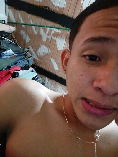 The image depicts an indoor scene within the city of Soacha, Colombia. A single individual, appearing male, is positioned in a close-up, chest-up shot. The person has light brown skin, short dark hair, dark eyes, and a subtle smile with slightly parted lips. A small silver-colored stud earring is visible in the left earlobe, and a thin, light-colored chain necklace is worn around the neck. The individual's left shoulder and upper chest are exposed. In the background, to the left of the individual, is a pile of folded clothing or textiles in various colors including red, blue, green, white, and black. The wall behind the subject is constructed of vertical light brown wooden planks featuring irregular white paint marks or patterns. Two dark horizontal beams and a thin green horizontal rod are visible traversing the background wall. The scene is lit consistently, suggesting an indoor environment.