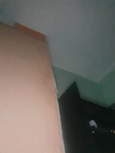 The image depicts an interior scene, likely a room, with a pale pink wall dominating the left side and extending towards the top. The wall appears to have some texture and slight discoloration. In the upper right portion of the image, a grey ceiling is visible, meeting the pink wall at an angle. A section of a darker, possibly green, wall or surface is present on the right, casting a shadow in the lower right quadrant. Within this shadow, a dark object, potentially furniture, is partially visible, with a small, bright white and orange reflection or object on its surface. The overall lighting is dim, and the image quality is somewhat blurry, suggesting it may have been taken with low light or a moving camera. The scene is devoid of any individuals. The location in Liubakhy, Ukraine, is not visually discernible from the provided image.