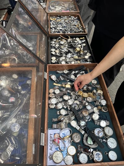 The scene depicts a vendor's stall at an outdoor market in Shinagawa, Japan. Multiple wooden display units are presented, filled with a large collection of timepieces. On the left, two multi-tiered wooden cases with clear glass covers are densely packed with various wristwatches, featuring both metal bracelets and leather straps. On the right, three open wooden trays are visible. The top two trays are rectangular and filled primarily with metal-banded wristwatches. The largest, lowest tray, lined with a blue-green fabric, contains a diverse assortment of wristwatches, several round pocket watches (some with attached chains), and loose watch components.

A person's right arm, covered by a dark short-sleeved garment, extends from the upper right of the frame, with their hand reaching into the lowest open tray. The index finger and thumb are actively manipulating or selecting an item from among the watches. Partially visible in this tray is a white rectangular card displaying "WE ACCEPT" alongside partial credit card logos (Visa, Mastercard). The ground beneath the stall is a light grey, consistent with pavement or concrete in an outdoor setting.