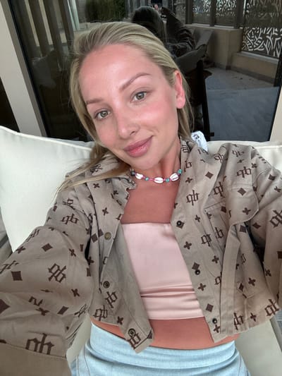 A fair-skinned blonde woman with green eyes is lying back on a white cushioned surface outdoors, likely a lounge chair or a settee. She is wearing a beige, patterned jacket over a pink top, a colorful shell necklace, and light-colored denim bottoms. The background shows an outdoor setting, possibly a balcony or a patio, with a dark window, and light patterned metal. The location is within the city of El Gouna, Egypt.