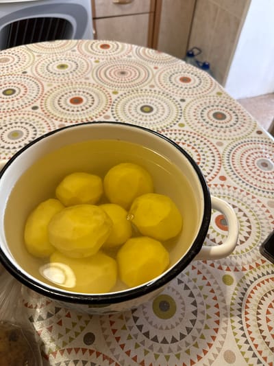 The image shows a white enamel pot filled with peeled potatoes submerged in clear liquid. The pot has black trim around the rim and a white handle. It is placed on a table covered with a tablecloth featuring a circular geometric pattern in shades of beige, green, orange, and gray. In the background, a kitchen counter with cabinets and a tiled backsplash is visible. The lighting suggests an indoor setting, likely a kitchen. The scene is devoid of any people or animals. The focus is on the pot of potatoes and the patterned tablecloth. The location is stated to be Морозовка, Russia.