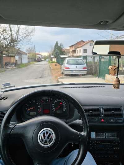 From the driver's seat of a Volkswagen, looking out onto a street in what appears to be Stara Reka, Bulgaria. The car's speedometer reads 1732. A silver car is parked ahead. The location is characterized by residential buildings, trees, and a narrow road. The steering wheel is prominently featured, with the Volkswagen logo clearly visible. The driver's leg is partially seen in the lower right. A wooden ornament hangs from the rearview mirror.
