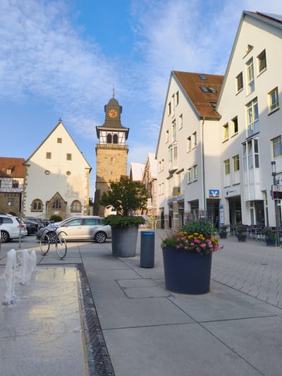The image shows a town square in Neuenstadt am Kocher, Germany, on a clear day with a blue sky and scattered clouds. In the background, a prominent clock tower with a red clock face stands beside a building with a white facade and a red-tiled roof. To the right, modern white buildings with numerous windows line the square. In the foreground, a fountain with jets of water is visible on the left, and a paved walkway leads towards the buildings. Several cars are parked on the side of the street. Flower pots with colorful flowers are placed along the walkway, and outdoor seating for a cafe is visible near the buildings on the right. A bicycle rack with two bikes is also present. The scene appears to be a peaceful urban setting.