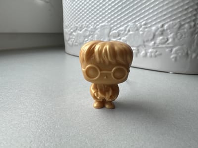 A close-up, eye-level shot shows a small, metallic gold-colored figurine of a character with round glasses and tousled hair, standing on a textured light gray surface. The figurine is positioned slightly off-center to the left. Behind the figurine, a white, textured object with a basket-like pattern is visible, blurred due to the shallow depth of field. The background is out of focus and appears to be a light gray wall or surface. The lighting suggests a bright, indoor environment. There is no discernible location in the city of Racibórz, Poland.