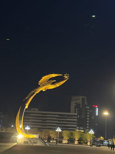 This nighttime image captures an illuminated urban scene, likely a public square or plaza in Shenhe District, China. The dominant subject is a grand, golden statue resembling a mythological bird, possibly a phoenix, with large, spread wings holding or emerging from a sun-like orb at its apex. The statue's base elegantly curves, illuminated from below, suggesting it's a significant public monument.

In the background, a multi-story building, likely a hotel or commercial establishment, stands tall with distinct striped patterns on its facade and some illuminated windows. Prominently displayed on its upper part is a red neon sign that reads "Hampton," accompanied by a series of bright white square lights below it. A circular street lamp casts a strong glow near the building.

The ground level features paved areas, indicative of a plaza or street, lined with decorative streetlights that have multiple globes. Trees with sparse foliage are also visible, some adorned with lights. On the far right, two indistinct figures appear to be walking, suggesting routine evening activity. A large vehicle, possibly a truck or bus, is partially visible near the building. The sky is dark, signifying late evening or night, and the scene is primarily lit by artificial sources, creating a vibrant urban ambiance.