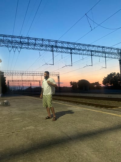 A man stands on a train platform at dusk in Samur, Russia, with overhead power lines and railway tracks visible. The man, wearing a white t-shirt and green patterned shorts, carries a white backpack. The sky displays a gradient of blue and orange hues. The platform is made of concrete, with yellow markings along the edge. Trees and distant structures are silhouetted against the colorful sunset.