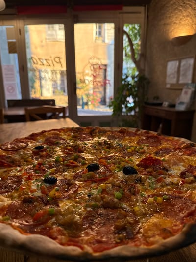 The image shows a close-up of a pizza with various toppings, including pepperoni, olives, and bell peppers, on a wooden table. In the background, through a glass door, the sunlit facade of buildings in Moustiers-Sainte-Marie, France, is visible. The text "Pizza" is reflected on the glass door, appearing backward. The scene appears to be indoors at a restaurant.