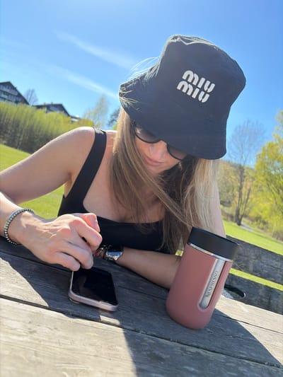 A woman is angled toward the camera, wearing a black "Miu Miu" bucket hat and sunglasses. She is looking at her phone which is resting on a grey wooden picnic table. A pink Nespresso thermos is also on the table, to her right. She is wearing a black top and a bracelet on her left wrist. A watch can be seen on her left arm. The photo was taken outdoors on a sunny day with a blue sky in Nærsnes, Norway. Green grass and trees are in the background.