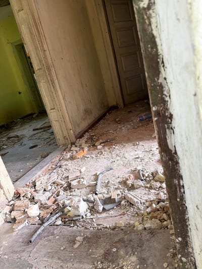 The image depicts an interior scene of severe decay and disrepair, possibly taken in Umarevtsi, Bulgaria, although no specific location is visible. The viewpoint is from above, looking down into what appears to be a room or section of a building. A portion of a painted door frame and the top of a door are visible in the upper left quadrant. The adjacent area reveals a wall covered in debris, including chunks of concrete, brick fragments, and plaster. Electrical conduit snakes across the surface of the debris. The wall itself shows signs of extensive damage, including crumbling plaster and exposed substrate. Below, the remnants of a yellow-painted wall is visible. An empty plastic bottle is stuck to the wall.