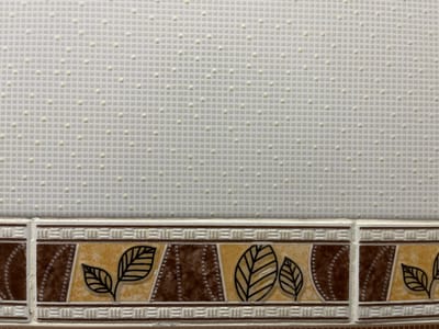 The image shows a close-up view of a textured white wall with a decorative tile border along the bottom. The white portion of the wall features a subtle grid pattern with raised dots, creating a tactile surface. The tile border is divided into repeating panels, each containing a design of stylized leaves and geometric patterns in shades of brown, beige, and white. The overall impression is of a clean, decorative surface, possibly a bathroom or kitchen wall. The image does not provide any information about its location in Quezon City, Philippines, nor does it contain any people, actions, or interactions.