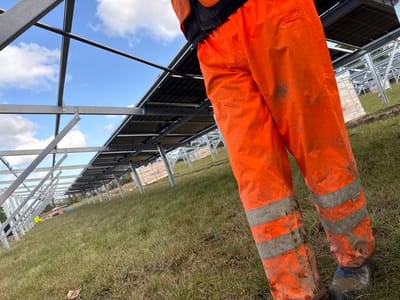 The image captures an outdoor construction site for a solar farm in Baldurgan Hill, Ireland. In the foreground, the lower body of an individual, from the mid-torso down, is visible, facing away from the camera. This individual wears soiled orange high-visibility work trousers with two horizontal silver-grey reflective stripes on each leg, and dark, muddy footwear. A black garment is visible beneath an orange upper-body garment. The individual stands on a grassy field with patches of dry or sparse vegetation.
Extending into the middle ground and background are numerous parallel rows of partially installed solar panel arrays. These arrays consist of galvanized metallic frameworks, some of which are fully constructed and hold multiple black photovoltaic panels. Other frameworks appear to be incomplete structures awaiting panel installation. The sky above is clear blue with scattered white cumulus clouds. In the distant background, wooden crates, some wrapped in white protective material, are visible; one crate bears the "JINKO" brand label. Additional metallic frameworks and small, brightly colored objects are visible on the ground beneath the structures further down the field.