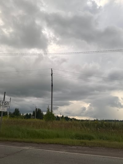 The image shows a roadside scene in Novoe Mart'yanovo, Russia, under a cloudy sky. In the foreground, a portion of a gray asphalt road is visible, with a white line on the left. To the right of the road, a dense, green, and brown grassy field stretches into the distance, bordered by a line of trees on the horizon. A tall wooden utility pole with multiple wires extending from it stands in the middle ground, positioned within the field. To the left of the pole, a white sign with black lettering is mounted on a gray pole, with the word "СТОП" (STOP) clearly visible. The sky is filled with dark, dramatic gray clouds, suggesting an overcast or stormy day.