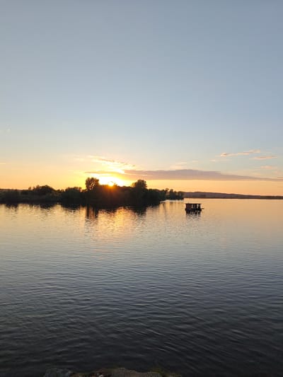 The image captures a serene landscape in Nikolo-Beryozovka, Russia, at either sunrise or sunset. A large body of water, likely a lake or wide river, occupies the majority of the frame, exhibiting subtle ripples and reflecting the warm hues of the sky. In the mid-ground, a dense line of trees and vegetation forms a dark silhouette across the horizon, with the sun partially obscured behind them, casting bright orange and yellow light and distinct sunbeams through the foliage. To the right of the center, a small, dark, rectangular floating structure, resembling a shed or small houseboat, rests on the water. The sky transitions from a brilliant orange and yellow near the horizon to a clear, pale blue overhead, with a few wispy clouds present, primarily on the right side. The far right horizon reveals a distant, hazy landmass. The immediate foreground shows a narrow strip of dark, rocky or gravelly ground, indicating the photo was taken from the shore. No people are visible.