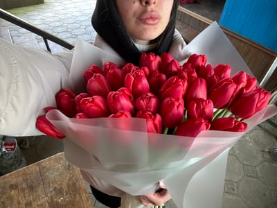 A female subject, visible from the chest up, is holding a large bouquet of red tulips in Trudobelikovskiy, Russia. She wears a light-colored jacket and a dark head covering, with her prominent lips visible in the upper frame. The bouquet consists of approximately 30-40 vibrant red, tightly closed or semi-closed tulips, wrapped in translucent white or light-pink paper. The subject is presenting the bouquet towards the camera. The flooring consists of light grey hexagonal or octagonal tiles with dark grout. To the lower left, a dark wooden surface is visible, alongside a clear plastic bottle with a red label resting in a dark, mesh-like container. In the background, a blue-painted wooden structure with vertical planks is present to the right, and a light-colored structure with a dark horizontal bar is visible in the upper left.
