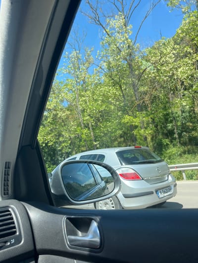 The image shows a view from inside a car. A silver car is visible through the side mirror. The license plate number on the silver car is A 5620 BB. The surrounding environment consists of trees and a blue sky, suggesting a rural or suburban setting. The presence of a car suggests the location is near a road. The location in the city of Zvezditsa, Bulgaria cannot be confirmed as it is not visible from the image.