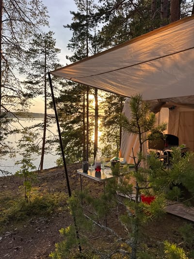 The image depicts a campsite on the shore of a lake at sunset, likely in the vicinity of Priladozhskoe, Russia, given the pine trees and calm water. A large tarp, supported by poles, forms a shelter over a small table. On the table, there is a bottle of red liquid, two glasses, and a mug. Behind the table, a portable grill and a tent are visible within the shelter. The ground is covered with pine needles and moss. The setting sun casts a warm glow through the trees, illuminating the scene and reflecting on the water. The overall impression is one of a tranquil camping experience in a natural environment.