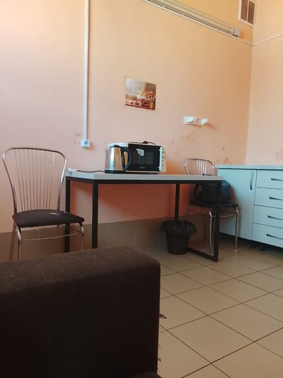 The image depicts a small room, possibly a communal area or kitchenette, within the Цімкавіцкі сельскі Савет region of Belarus. The walls are painted a light peach color. In the foreground, a dark brown, upholstered sofa is partially visible, occupying the lower left portion of the frame.
The central focus of the room is a black-framed table with a white top, positioned against a peach-colored wall. On the table, a silver electric kettle with a black handle and a black microwave oven are placed. A black cable extends from the kettle. To the left of the table, a chrome-framed chair with a dark seat is visible. To the right of the table, another chrome-framed chair with a dark seat is occupied by a dark blue backpack. Beneath the table, a black trash can with a plastic liner is present.
Further to the right, a white cabinet with multiple drawers and silver handles is situated against the wall. On the peach wall above the cabinet, a small framed picture featuring food items is displayed. A white electrical conduit runs vertically along the wall to the left of the table, with a white electrical outlet and a "220V" label near its base. A white double-cup holder fixture is mounted on the wall to the right of the picture. Fluorescent lighting fixtures are visible along the ceiling line. The floor is covered with beige square tiles. The overall impression is one of a functional, sparsely furnished space.