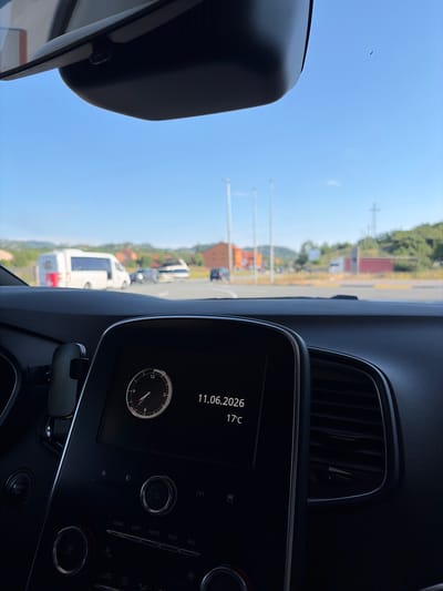 The image, taken from the interior of a car, shows a dashboard display with a clock and the date 11.06.2026, along with a temperature reading of 17°C. Outside the car, through the windshield, a street scene unfolds under a clear blue sky. Several vehicles are visible, including a white van with red markings, and other cars. Buildings and hills with vegetation form the background. The scene appears to be in Očinići, Montenegro, based on the context provided.