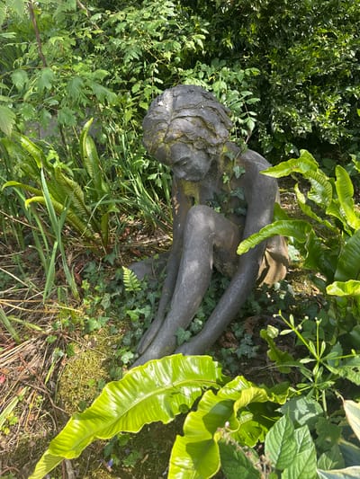 The image shows a weathered, dark gray statue of a child sitting with its head bowed, partially covered in moss and ivy. The statue is surrounded by lush green foliage, including ferns and various leafy plants, in what appears to be a garden setting. The lighting suggests it is daytime, with sunlight filtering through the leaves, casting shadows. The statue itself is centrally positioned, appearing aged and integrated into the natural environment. There are no people present in the image. The scene is located in Mid Sussex, United Kingdom.