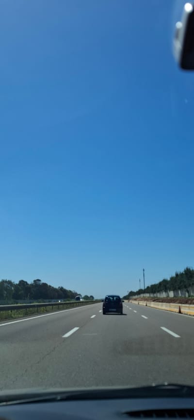 The image depicts a road, possibly a highway, with vehicles traveling on it. The view is from inside a car, looking out onto the road. The sky is clear and blue. Trees line the side of the road. The location is specified as Harhoura, Morocco, but there are no visual cues in the image that definitively confirm this specific location.
