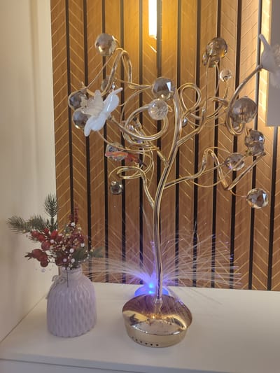 The image depicts a still life arrangement on a white shelf against a wooden paneled wall with alternating dark and light wood grain patterns. In the foreground, to the left, is a pale lavender ceramic vase with a textured surface and a small fabric bow tied around its neck. The vase contains a small arrangement of artificial greenery, red berries, and white beads, resembling a festive decoration. To the right of the vase is a decorative lamp with a gold-colored base and a central stem from which metallic branches extend. These branches are adorned with numerous clear, faceted crystal-like ornaments and a white floral embellishment. The base of the lamp emits a soft blue light, and thin, white fiber optic strands radiate outwards from it, creating a luminous effect. The background features a vertically oriented wooden paneling with a chevron pattern. A bright, vertical light fixture is visible in the upper portion of the image, casting warm illumination on the scene. There are no people or animals present in the image. The location is not specified in the image or provided by the user.