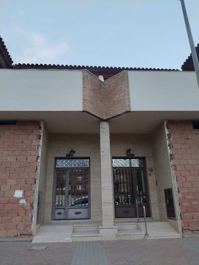 The image shows the front of a building with two separate entrances, each with a metal door and steps leading up to it. The facade is a mix of brick and what appears to be a light-colored stone or plaster. A central pillar supports the roof, which has a distinctive decorative brick pattern above a white section. The building's exterior looks somewhat unfinished, with exposed brickwork on the sides.