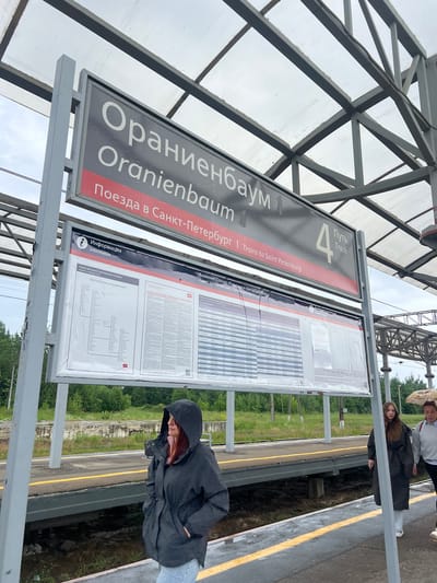 The image captures a train station platform in Lomonosov, Russia. A large sign displays "ОРАНИЕНБАУМ 1" (Oranienbaum 1) in white Cyrillic letters, with "Oranienbaum" in English below it. A red band runs across the sign, with "Поезда в Санкт-Петербург 1" (Trains to Saint Petersburg 1) in Cyrillic and "Trains to Saint Petersburg" in English. To the right, a large number "4" is prominently displayed, with "Путь" (Path) and "Track" written below it in Cyrillic and English respectively.

Below this, a large information board details train schedules and routes. The board is covered by a protective clear plastic sheet. A diagram showing "Балтийское направление" (Baltic direction) of suburban trains is visible on the left side of the board.

Three individuals are present on the platform. The closest is a person wearing a dark hooded jacket and light-colored pants, standing with their hands in their pockets, looking to the right. Further down the platform, two individuals are walking away from the camera. One, in a dark coat, is carrying a light-colored umbrella.

The platform is a concrete structure with yellow safety lines along the edge. The background consists of trees and greenery, suggesting a semi-urban or suburban setting. The station is covered by a white translucent roof supported by a metal frame. The sky is overcast.