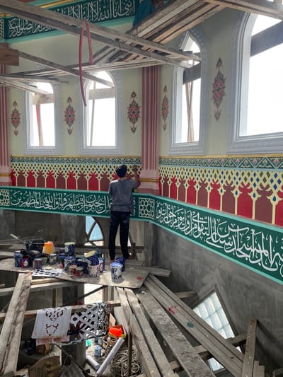 The interior of a building, likely a mosque, in Mns Asan, Indonesia, is depicted under active decoration. A male individual, seen from behind, stands on a wooden platform, painting intricate patterns onto a wall section with a brush. He wears a grey shirt and dark trousers. Numerous paint cans, brushes, and stencils are arranged on a wooden platform to his left. A second individual is partially visible on higher wooden scaffolding in the upper right background.
The walls feature elaborate Islamic calligraphy in white script on a prominent green band, extending horizontally across the room. Above this, a decorative frieze combines red, gold, and white geometric and stylized floral motifs. Pale yellow walls ascend to the ceiling, punctuated by multiple tall, arched windows that are currently open, allowing bright daylight to enter. Pinkish-red pilasters or columns flank some window sections, which are also adorned with smaller, intricate decorative patterns. Extensive temporary wooden scaffolding structures crisscross the space, supporting both workers and materials. The lower wall section below the finished decoration is unfinished grey concrete. A smaller, arched window with a glass block infill is visible in the lower right background. Overhead, a red flexible tube hangs from the wooden ceiling structure.