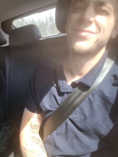 A man is sitting in the passenger seat of a car, looking at the camera. He is wearing a dark blue polo shirt and a seatbelt. The interior of the car is visible, including the headrest and the seat. Through the window, a forest and sky can be seen. The man has tattoos on his arm, including a skull and crossbones and an owl. The image appears to be taken during the daytime, with sunlight streaming through the window, creating bright highlights and shadows. The location is specified as Tolokonnikovo, Russia.