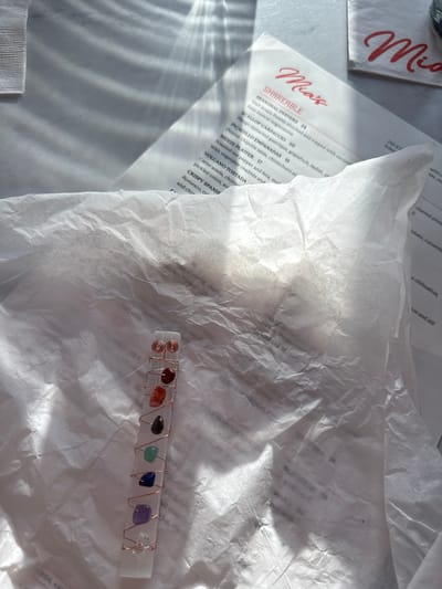 This image captures a tabletop scene, likely in a restaurant in Solana Beach, United States, indicated by visible text on the menu. The main subjects are a white menu from "Mia's" restaurant, a crumpled piece of white paper (possibly parchment or tissue paper) partially obscuring the menu and table, and a decorative object resembling a selenite wand adorned with various colored gemstones wrapped in copper wire.
The activity taking place is implied to be dining or waiting to order at "Mia's" restaurant. The presence of the menu suggests a mealtime setting. The crystal object appears to be a personal item or a decorative piece associated with the setting.
Notable details include the bright, natural light casting distinct shadows and highlights across the scene, suggesting it is daytime. The light creates a textured appearance on the crumpled paper and reflects off the shiny surfaces of the crystals. The object features a clear, rod-like crystal (likely selenite) with seven small, polished gemstones of different colors (red, orange, dark, green, blue, purple, and clear/white), sequentially arranged and secured with coiled copper wire, reminiscent of chakra colors.
Visible text includes the prominent "Mia's" logo on the menu and a napkin/coaster. The menu lists "SHAREABLE" items such as "SEASONAL OYSTERS 24", "SCALLOP CARPACCIO 20", "PICADILLO EMPANADAS 15", "MEZZE PLATTER 17", "VOLCANO TOSTADA", and "CRISPY SPANIS". Ingredients and descriptions like "Half dozen freshly shucked and topped with cucum", "lime mezcal mignonette", "Mango tamarind gastrique, grapefruit, radish, sh", "Peanut chipotle salsa, chimichurri ai", "with vegetable crudités and", "Roasted red pepper and feta, mu", "Carne asada, chorize", "pickled onion, av", "Romesco, cr", and "and chir" are also partially legible. Fragments of an address, "243 N" and "Solana", further specify the location context. Other obscured words like "o chihuahua", "on and dill", "charred pinea", and "crouton topped with" are also discernible through the paper.