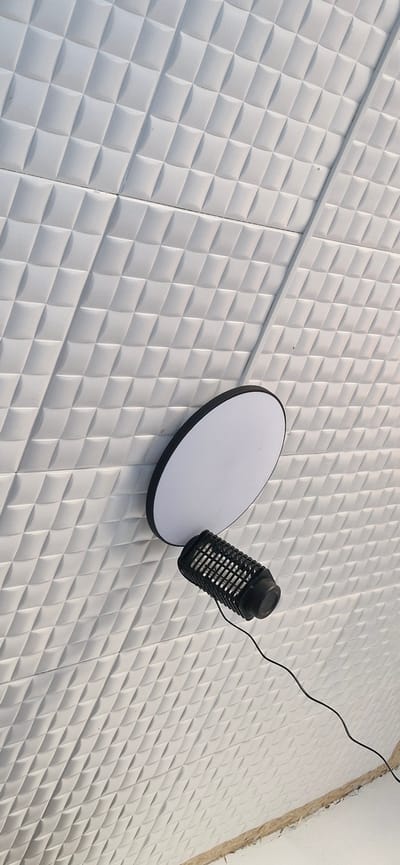 A black mosquito repellent device with a wire is positioned in front of a circular white light fixture, both mounted on a textured white wall. The wall features a repeating pattern of raised diamond shapes. The image captures a ceiling or wall in an interior space.
