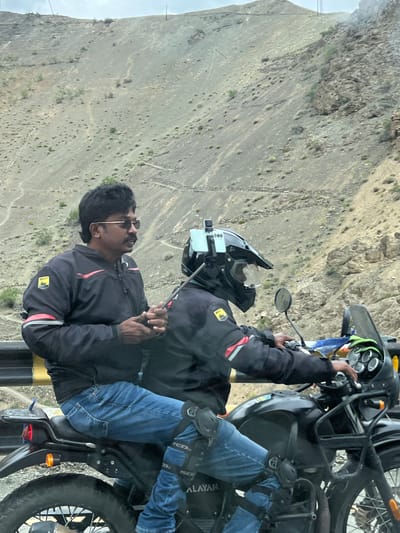 Two individuals are riding a black Royal Enfield motorcycle through a mountainous, arid landscape. The rider in front is wearing a black motorcycle helmet with a camera mounted on top, a black jacket, and blue jeans. The passenger behind the rider is wearing sunglasses, a black motorcycle jacket with red accents, and blue jeans. They appear to be on a road or trail in a mountainous region, with a barren, rocky hillside as the backdrop. The sky is overcast. The image quality is moderate, with some blur and noise. There are no identifiable landmarks or signs of civilization that would indicate a specific location within Lotsum, India.