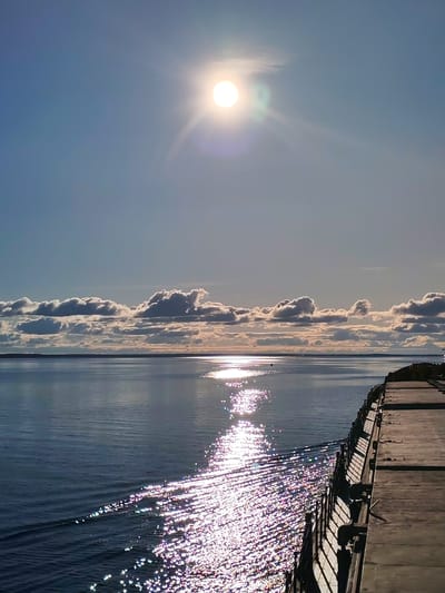 The photograph presents a view of a body of water, likely a sea or large lake, under a bright, sunny sky with scattered clouds. The sun is high in the sky, its light reflecting off the water's surface, creating a sparkling effect. A dock or pier extends into the water from the right side of the frame. In the distance, a strip of land is visible on the horizon, and above it, a band of clouds stretches across the sky. No people are visible in the scene. The image evokes a sense of calmness and vastness, with the interplay of light and water being a dominant feature. I don't know if it is taken in the city of Vichelovo, Russia.