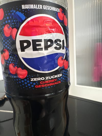 The media file presents a close-up, sideways view of a Pepsi Zero Sugar Cherry beverage bottle, likely placed indoors. The bottle is predominantly black, featuring a prominent label with the iconic red, white, and blue Pepsi logo at its center. Surrounding the logo and across the label's black background are illustrations of vibrant red cherries, complemented by small blue dots.
Visible text on the label is in German, reflecting the location context of Fulda, Germany. In large letters, the brand name "PEPSI" is clearly displayed. To the left of the logo, white text reads "MAXIMALER GESCHMACK," which translates to "MAXIMUM TASTE." To the right, "ZERO ZUCKER" (ZERO SUGAR) is written in white, followed by "CHERRY GESCHMACK" (CHERRY TASTE) in red letters.
The background is partially blurred, revealing horizontal lines that suggest window blinds or a textured window, along with a white frame. This indicates an indoor setting, possibly near a window. The surface on which the bottle rests appears dark. The image is a static product shot with no discernible activity or specific time of day.