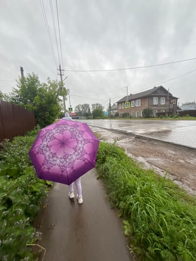 The image depicts a person walking with a purple umbrella on a wet sidewalk in Balezino, Russia. The person, seen from behind, is wearing light-colored pants and white shoes. The umbrella, held aloft, has a decorative pattern. To the left of the sidewalk is a brown wooden fence and lush green vegetation. Across the street, a two-story wooden house with many windows stands under an overcast sky. The road surface is wet and shows signs of wear, with puddles reflecting the dull light. A pedestrian crossing is visible on the road. Power lines are strung overhead, supported by utility poles. The overall atmosphere is that of a rainy or recently rained-upon day in a semi-rural or suburban setting.